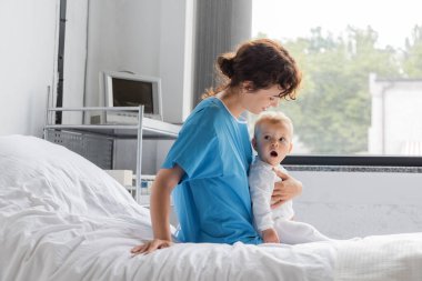 baby girl with open mouth looking away near mother in patient gown sitting on hospital bed
