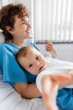 happy woman smiling on hospital bed near baby looking at camera on blurred foreground