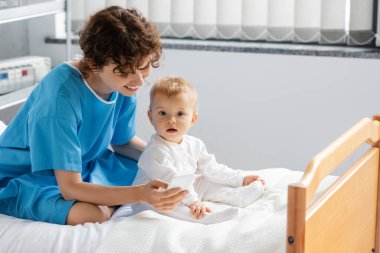 smiling woman in patient gown using smartphone near baby in romper sitting on hospital bed clipart