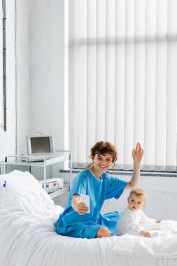 smiling woman waving hand during video call on smartphone near baby girl on hospital bed 
