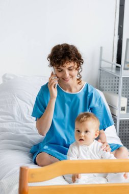 smiling woman with closed eyes talking on smartphone near little baby on hospital bed