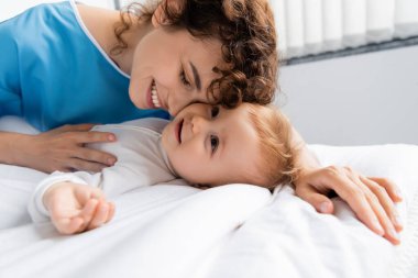smiling woman in patient gown embracing toddler child lying on bed in hospital ward