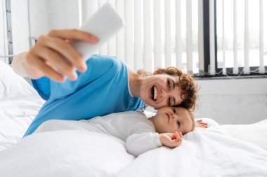 excited woman with mobile phone taking selfie with toddler child on bed in hospital ward clipart