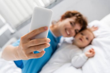 selective focus of mobile phone in hand of happy woman taking selfie with little daughter in clinic clipart