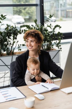 joyful businesswoman looking away while sitting with baby at work desk near blank notebook and blurred coffee cup clipart