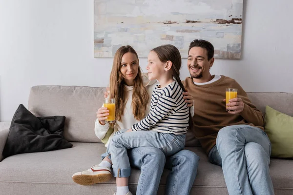 cheerful daughter sitting on laps of mother near positive father holding glass of orange juice 