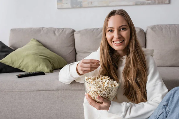 stock image cheerful woman holding bowl with popcorn while watching movie in living room