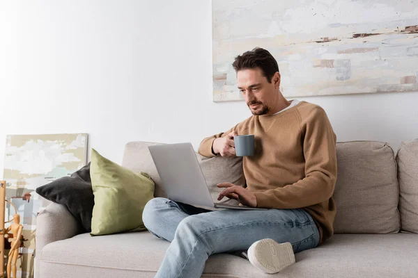 bearded man using laptop while holding cup and sitting on sofa in living room