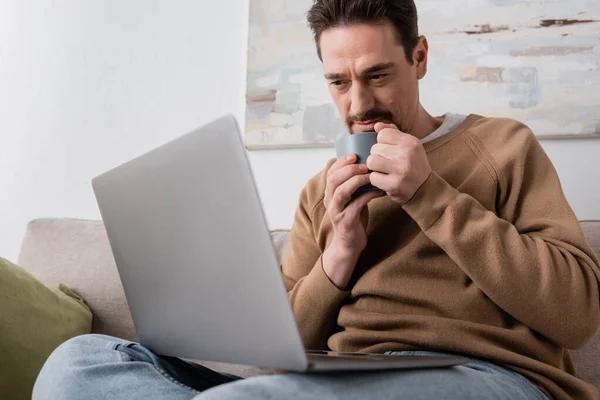 bearded man using laptop while holding cup and sitting on couch in living room