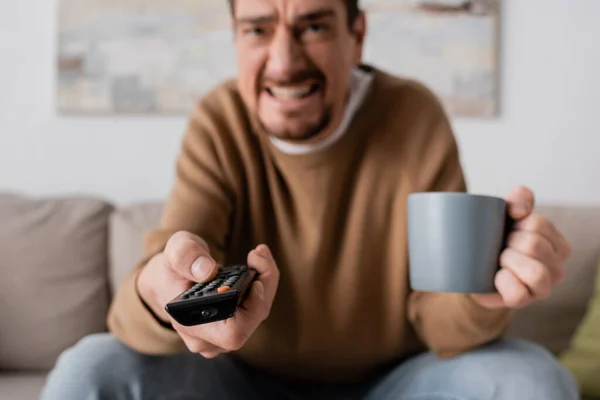 angry man holding remote controller and cup of coffee in living room