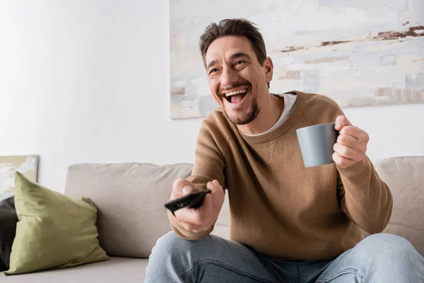 amazed man holding remote controller and cup of coffee in living room