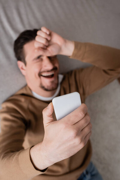 вид сверху на бородатого мужчину в бежевом свитере, держащего смартфон и опирающегося на диван 