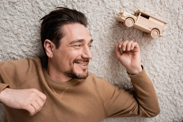 top view of bearded and cheerful man lying on carpet near wooden toy car in living room 
