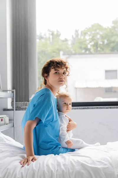 woman in patient gown looking at camera while sitting with little daughter on hospital bed near window
