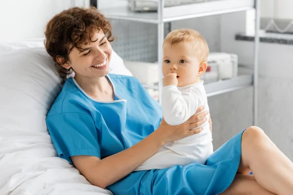 cheerful woman in hospital gown looking at toddler daughter holding hand near mouth and looking away in hospital ward