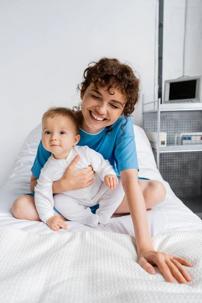 excited woman laughing with closed eyes and hugging baby on bed in hospital ward