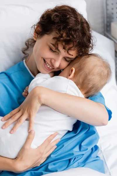 happy woman with closed eyes embracing little child in romper on bed in hospital