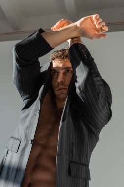 shirtless man in black and striped blazer posing with crossed arms above head on grey background