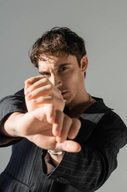 trendy brunette man in black blazer posing with outstretched hands while looking at camera on blurred foreground isolated on grey