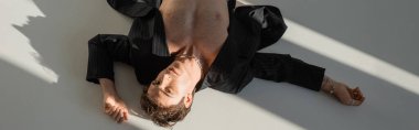 top view of shirtless man in black blazer and silver necklace lying and looking at camera on grey background, banner