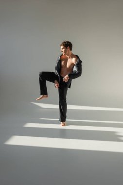full length of barefoot man in black trousers and blazer on shirtless body standing on one leg on grey background