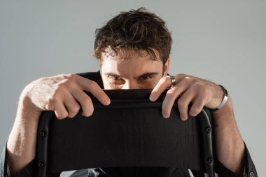 man in silver bracelet and finger ring hiding behind chair back while looking at camera isolated on grey