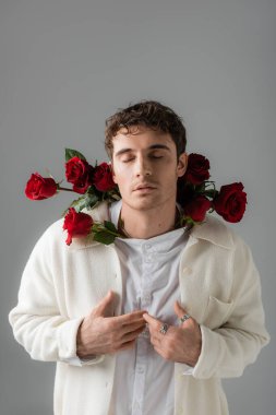 brunette man with closed eyes posing with red roses in white jacket and shirt isolated on grey