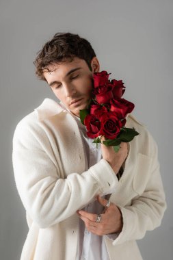 stylish brunette man with closed eyes holding bouquet of red fresh roses isolated on grey