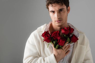 front view of man in white trendy clothes looking at camera while standing with bouquet of red roses isolated on grey