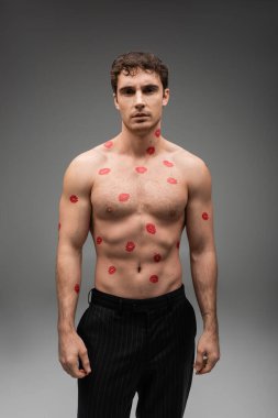 front view of sexy shirtless man with red kiss prints on body looking at camera on grey background clipart