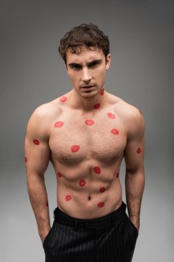 shirtless man with red kisses on muscular body standing with hands in pockets of black pants on grey background