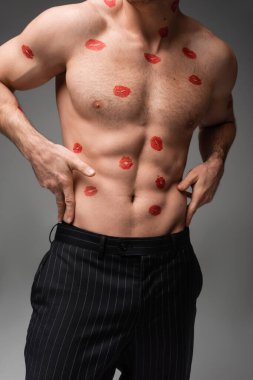 cropped view of man in black pants touching muscular torso with red lip prints isolated on grey