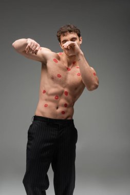 athletic man with red kisses on shirtless body obscuring face while posing with closed eyes on grey background