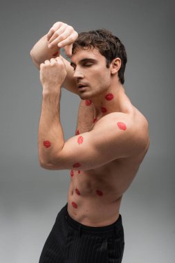 shirtless muscular man with red kiss prints on body posing with closed eyes and hands near face on grey background clipart