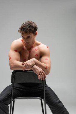 brunette muscular man with red kiss prints on body sitting on chair isolated on grey