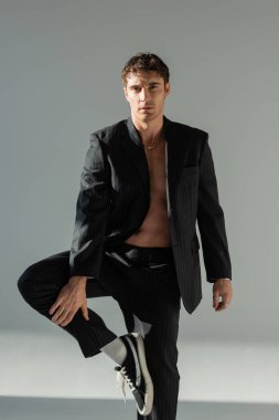 sexy brunette man in black pants and blazer on shirtless body standing on one leg and looking at camera on grey background