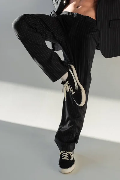 cropped view of trendy man in black trousers and sneakers posing on one leg on grey background