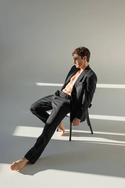 full length of relaxed barefoot man with muscular torso posing in black suit while sitting on chair on grey background