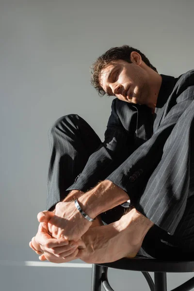 low angle view of barefoot man with closed eyes sitting in black suit and silver bracelet on grey background