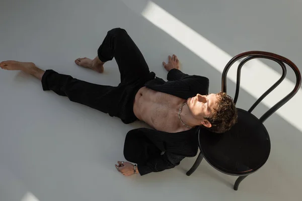 top view of sexy barefoot man with muscular torso lying in black suit near chair on grey background