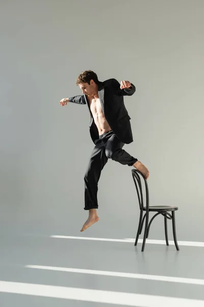 full length of muscular man in black suit jumping from chair with outstretched hands on grey background