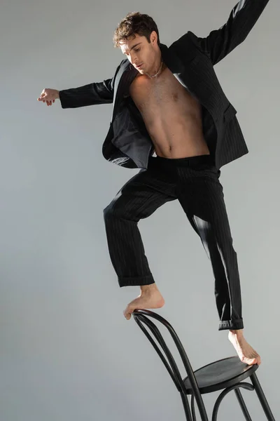 full length of barefoot man in black suit on shirtless body balancing on chair isolated on grey