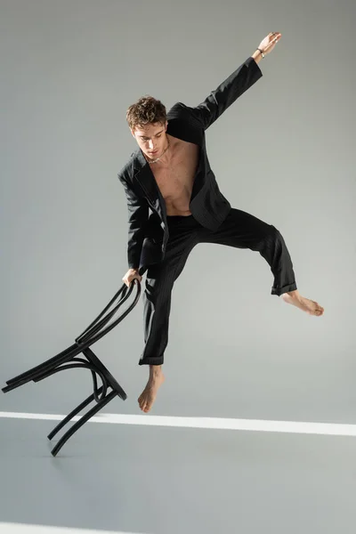 full length of barefoot man in black stylish suit doing trick while jumping with chair on grey background
