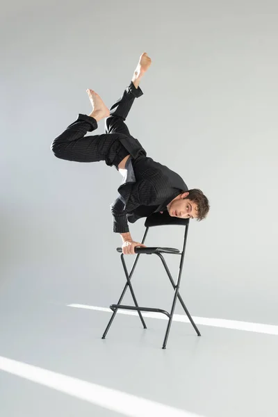 full length of stylish barefoot man in black suit looking at camera while doing trick on chair on grey background