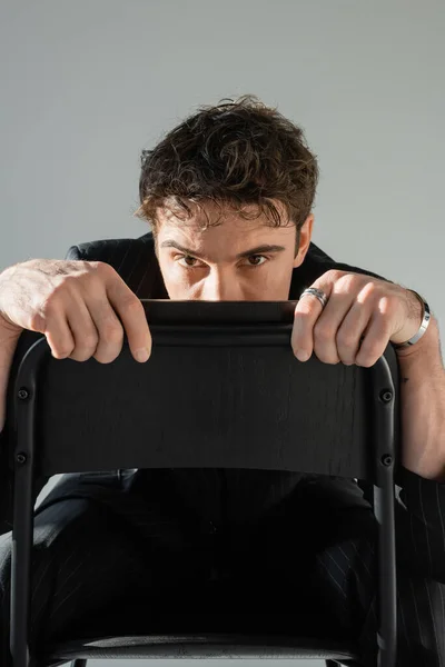 stock image brunette man in black clothes hiding face behind chair back and looking at camera isolated on grey