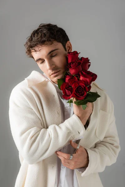 stock image stylish brunette man with closed eyes holding bouquet of red fresh roses isolated on grey