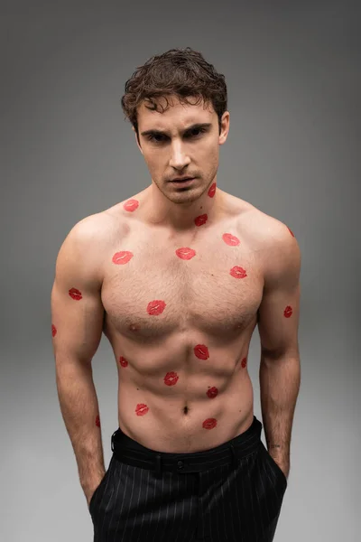 stock image shirtless man with red kisses on muscular body standing with hands in pockets of black pants on grey background