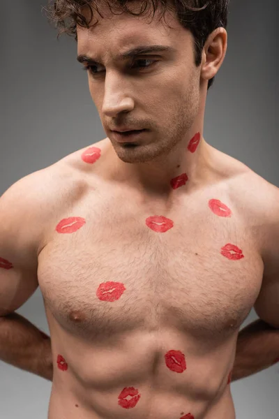 shirtless man with red kisses on muscular body standing with hands behind back on grey background