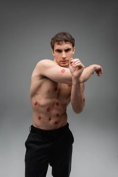 sexy shirtless man with red lipstick prints on body looking at camera while standing on grey background
