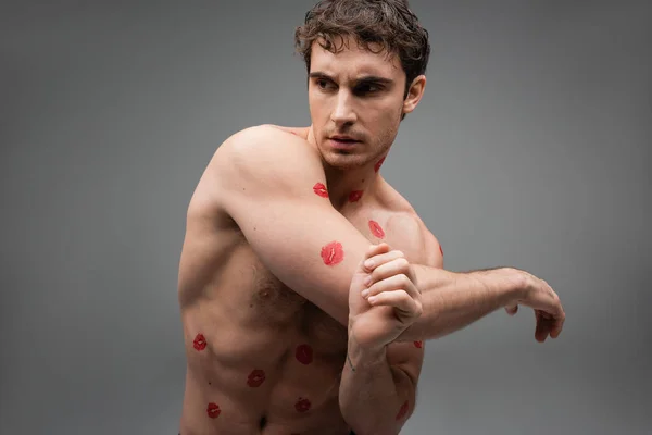 shirtless and muscular man with red lipstick marks on body looking away isolated on grey